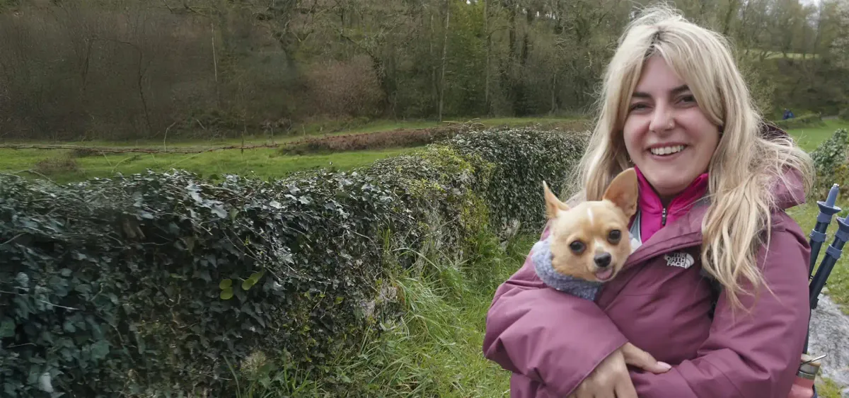Peregrina con chaqueta morada sosteniendo a su perro en brazos, rodeada de naturaleza y vegetación, disfrutando de una etapa tranquila del Camino de Santiago.