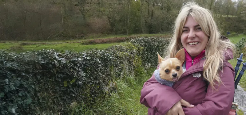 Peregrina con chaqueta morada sosteniendo a su perro en brazos, rodeada de naturaleza y vegetación, disfrutando de una etapa tranquila del Camino de Santiago.