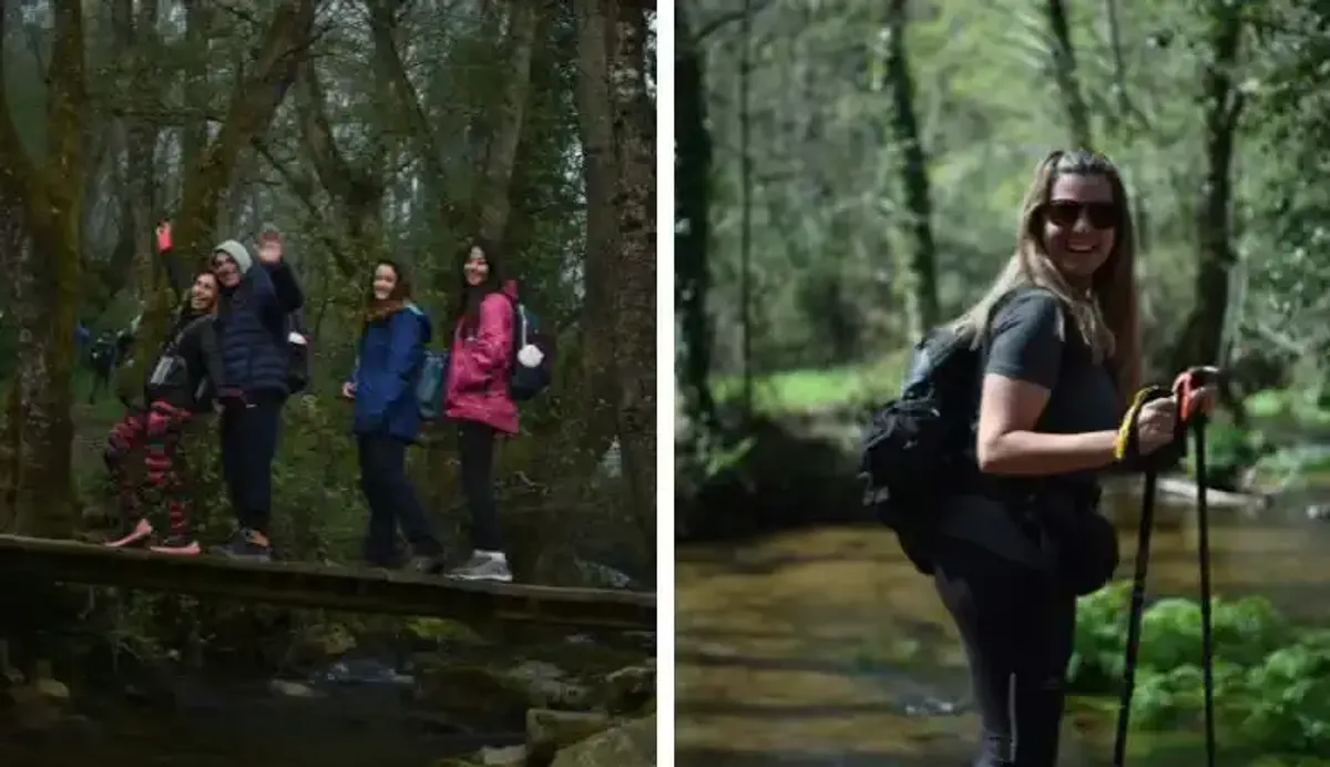 Senderistas sonriendo cruzan un puente sobre un río. Con mochilas y bastones, disfrutan de su caminata grupal por un tramo boscoso del Camino de Santiago.