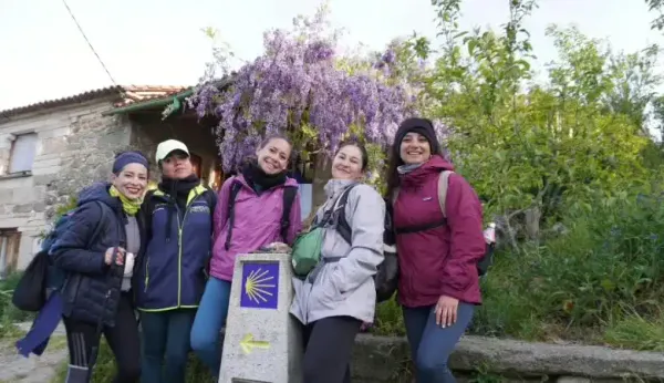 Un grupo de cinco excursionistas sonriendo posa junto a un mojón del Camino de Santiago, que muestra la icónica concha de vieira y una flecha amarilla direccional.