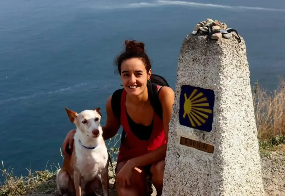 Una mujer sonríe junto a su perro frente al mojón de 0,00 km del Camino de Santiago, con el mar de fondo y piedras colocadas sobre la señal del peregrinaje.