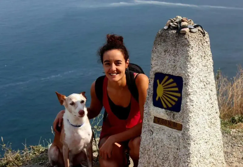 Una mujer sonríe junto a su perro frente al mojón de 0,00 km del Camino de Santiago, con el mar de fondo y piedras colocadas sobre la señal del peregrinaje.